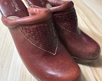 May include: A pair of brown leather clogs with a woven design on the front. The clogs have a wooden heel and are labeled "The Wild Pair" and "Size 38".