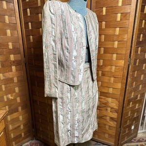 May include: A vintage two-piece suit featuring a jacket and a knee-length skirt. The suit is made of a light-colored fabric with vertical stripes and a floral pattern in pink and green. The jacket has long sleeves and a collarless design.