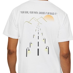 Puede incluir: Camiseta blanca con un diseño gráfico en la espalda. El diseño presenta una carretera con farolas, montañas y un sol. El texto "YOUR GOAL, YOUR PATH. CHOOSE IT OR BUILD IT." está impreso encima.
