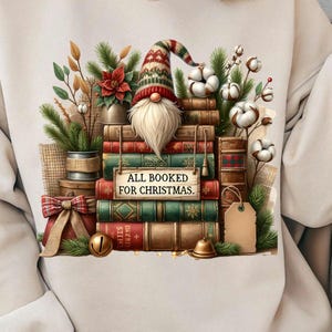 Peut inclure: Un sweat-shirt beige avec une illustration d'un gnome à longue barbe blanche et bonnet à motifs, posé sur une pile de livres anciens. Les livres rouges et verts portent une pancarte ALL BOOKED FOR CHRISTMAS, entourés de pin, de coton et d'un nœud.