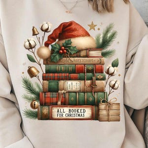 May include: A beige sweatshirt featuring a stack of books adorned with a Santa hat, holly, and Christmas decorations. The books are green, red, and brown, with the text "ALL BOOKED FOR CHRISTMAS" on a small sign.