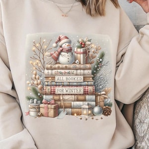 May include: Beige sweatshirt featuring a stack of books with a snowman wearing a hat and scarf. The books have the text "ALL BOOKED FOR WINTER". Presents, pinecones, and decorative lights surround the books, creating a cozy winter theme.