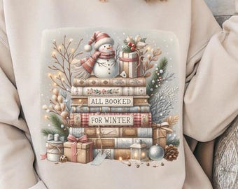 May include: A beige crewneck sweatshirt featuring a graphic print of a stack of books with the text ALL BOOKED FOR WINTER. The illustration includes a snowman wearing a scarf, wrapped gift boxes, pine branches, berries, and a lantern.