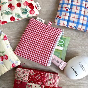 Può includere: Una collezione di piccole pochette in tessuto con vari motivi e colori. I disegni includono quadretti rossi, patchwork floreale e stampe di ciliegie. Una pochette è aperta, rivelando un tubetto di balsamo per le labbra e denaro.