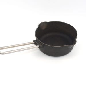 May include: A small, black cast iron saucepan with a curved rim and a silver-colored handle. The handle is attached to the side of the pan and curves upward. The pan is designed for cooking and is likely used for heating liquids or small portions.