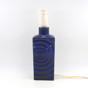 Könnte beinhalten: Ein rechteckiger Lampenfuß aus Keramik in tiefblauem Glanzfinish mit einem erhabenen konzentrischen Kreismuster auf der Vorderseite. Die Lampe verfügt über eine weiße Fassung an der Oberseite und ein helles Stromkabel, das seitlich herausführt.