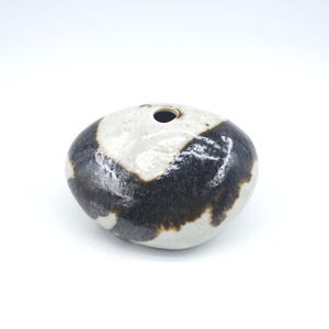 Puede incluir: Un pequeño jarrón de cerámica redondeado con una sola abertura. El jarrón presenta un diseño de dos tonos, con una combinación de colores blanco y negro. La sección negra tiene un aspecto moteado, y el jarrón está sobre un fondo blanco.