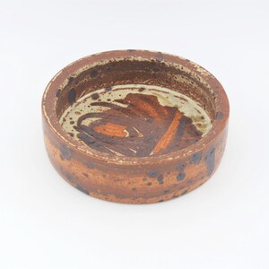 May include: A small, round ceramic bowl with a brown, white and orange glaze. The bowl has a speckled and mottled appearance.