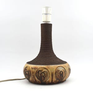 May include: A brown ceramic table lamp with a textured, vase-like body and a cream-colored base. The base features carved floral designs. The lamp has a standard light bulb socket and a power cord. The lamp measures approximately 12 inches tall.