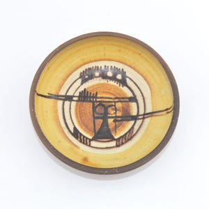 May include: A small, round ceramic bowl with a brown rim and a light yellow interior. The bowl features a central design with black and brown geometric shapes and lines. The bowl is likely handmade and suitable for serving small portions.