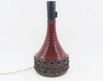 May include: A red ceramic table lamp with a textured base and a black lampshade. The base has a unique design with small holes around the bottom.