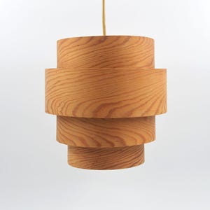 May include: A wooden pendant light fixture with a layered, cylindrical design. The light is made of light-colored wood and hangs from a thin, beige cord. The fixture has a stacked appearance, with each layer slightly smaller than the one below.