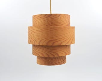May include: A wooden pendant light fixture with a layered, cylindrical design. The light is made of light-colored wood and hangs from a thin, beige cord. The fixture has a stacked appearance, with each layer slightly smaller than the one below.