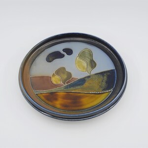 May include: A round ceramic plate with a blue rim and a landscape scene painted in shades of brown, green, and yellow. The scene depicts a hillside with two trees and a cloudy sky.