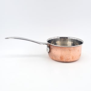 May include: A small saucepan featuring a polished copper exterior and a stainless steel interior. The pan has a long, slender, curved metal handle attached to the side with a single visible rivet, providing a functional grip for cooking tasks.