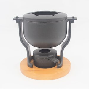 Könnte beinhalten: Ein schwarzer Fondue-Topf aus Gusseisen mit Holzsockel. Der Topf hat einen Deckel mit Öffnungen für Gabeln und steht auf einem Ständer mit einem Brenner. Der Holzsockel ist rund und hellbraun.