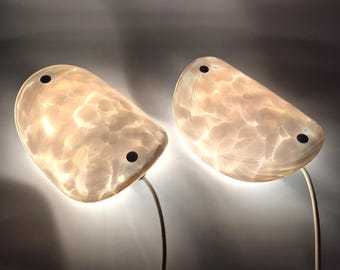 May include: Two white, semi-circular wall sconces with a cloudy, translucent glass shade. Each sconce has a black wire cord.