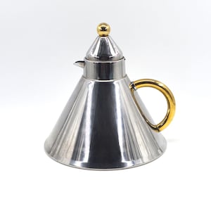 May include: A stainless steel teapot with a conical body and a gold-colored handle. The lid is topped with a small gold sphere. The teapot has a polished, reflective surface and a spout for pouring.