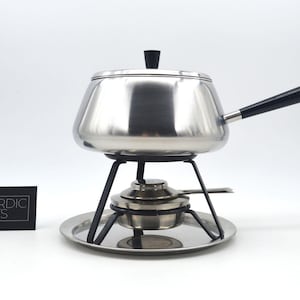 Può includere: Una fonduta in acciaio inossidabile con manico e pomello del coperchio neri, posta su un supporto in metallo nero con un bruciatore e una base rotonda. La fonduta è esposta con un biglietto nero con la scritta "NORDIC ARTS".