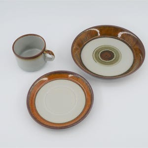 May include: A ceramic cup, saucer, and bowl set. The cup and saucer have a light grey interior and a brown rim. The bowl has a white centre with concentric green and brown circles. The set is on a white background.