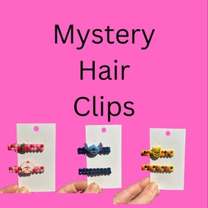 May include: The image shows three sets of hair clips against a pink backdrop, with the text "Mystery Hair Clips". Each set contains two clips. One set features a strawberry and a character clip. Another has a blue character clip and a beaded clip. The last set has a yellow character clip and a beaded clip.