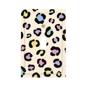 May include: A rectangular light switch wall plate featuring a cream-coloured background with a repeating leopard print pattern. The spots are outlined in black and filled with pastel colours including pink, yellow, light blue, purple, and mint green.