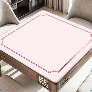 May include: A square mahjong table mat featuring a pale pink surface with a dark red double-line border that curves inward at each corner. The mat is designed to fit securely on top of a wooden table, providing a smooth playing area for games.