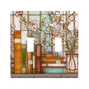 Op de afbeelding: Een decoratieve dubbele lichtschakelaar cover met een glas-in-lood ontwerp. De afbeelding toont stapels boeken, bloemen en een vaas met bladeren, allemaal in warme tinten oranje, bruin en groen. De achtergrond heeft een rasterpatroon.