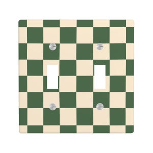May include: A decorative double light switch cover with a checkerboard pattern. The design features alternating squares of cream and dark green. Two light switch openings and four silver screws are visible.