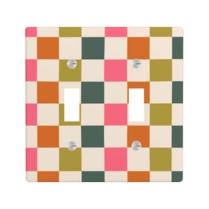 May include: A double light switch wall plate featuring a repeating checkered pattern in shades of pink, olive green, burnt orange, and cream. The square plate includes two rectangular cutouts for toggle switches and four silver metal screws.