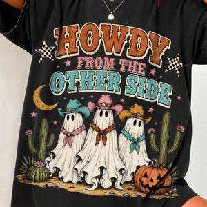 May include: A black short-sleeve t-shirt featuring a graphic print with the text "HOWDY FROM THE OTHER SIDE". The design includes three ghosts wearing cowboy hats and bandanas, two cacti, a crescent moon, stars, and a jack-o'-lantern pumpkin.