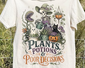 May include: An ivory short-sleeve t-shirt featuring a graphic of a skeleton in a witch hat watering plants, a black cat, a ghost, and a pumpkin. The text reads: PLANTS, POTIONS & POOR DECISIONS. The design includes vines, flowers, and a venus flytrap.