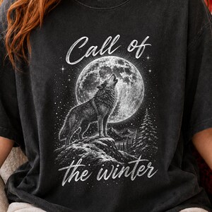 May include: A charcoal black t-shirt featuring a white graphic print of a wolf howling at a large full moon. The design includes a mountain landscape with pine trees and the text Call of the winter in a stylized script font across the center.
