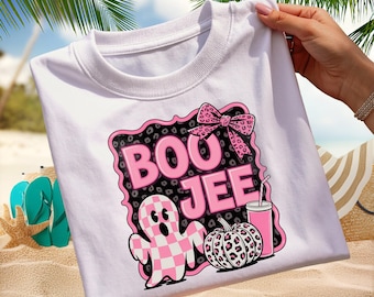 Może przedstawiać: Biała koszulka z różowo-czarną grafiką z napisem "BOO JEE". Grafika zawiera ducha, kokardę, dynię i napój. Duch ma różowo-biały wzór w szachownicę.