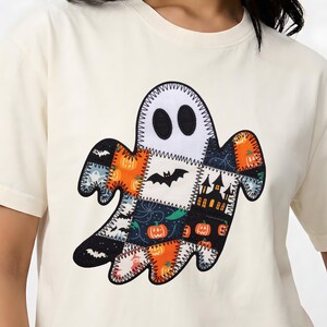 Może przedstawiać: Kremowy t-shirt z krótkim rękawem z dużą grafiką ducha na środku. Duch został zaprojektowany z patchworkowym wzorem z różnych tkanin o tematyce Halloween, w tym pomarańczowych dyń, czarnych nietoperzy i sylwetki nawiedzonego domu.