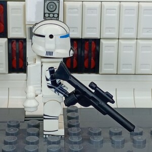 May include: A white and blue Lego minifigure of a clone trooper holding a black blaster rifle. The minifigure has a white helmet with blue accents and black markings. The background features white panels and gray Lego baseplates.