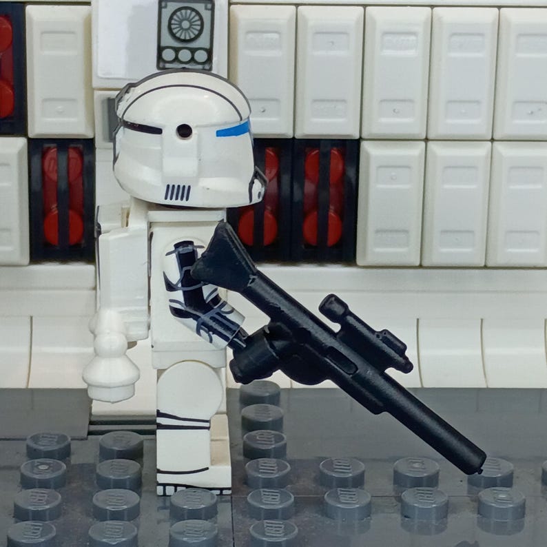 May include: A white and blue Lego minifigure of a clone trooper holding a black blaster rifle. The minifigure has a white helmet with blue accents and black markings. The background features white panels and gray Lego baseplates.