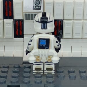 May include: A white and black Lego minifigure of a space trooper, viewed from the back. The figure has a white helmet and armor with black accents. A black weapon is attached to the right side. The background features white and red blocks.