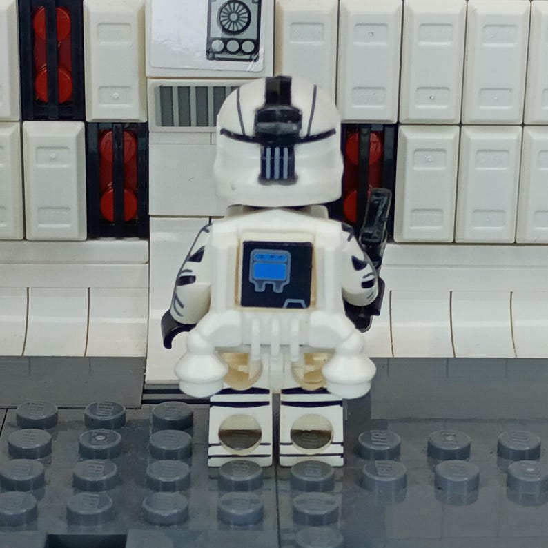 May include: A white and black Lego minifigure of a space trooper, viewed from the back. The figure has a white helmet and armor with black accents. A black weapon is attached to the right side. The background features white and red blocks.