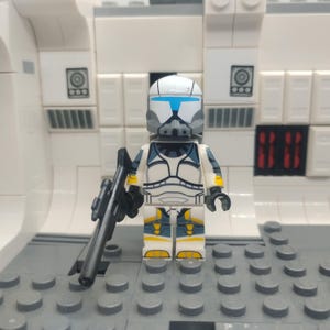 Könnte beinhalten: Eine Lego-Minifigur eines Soldaten in weißer, blauer und gelber Rüstung, der ein schwarzes Gewehr hält. Der Soldat hat einen weißen Helm mit blauen Akzenten und eine graue Gesichtsmaske. Der Hintergrund ist eine weiße und graue Lego-Struktur.