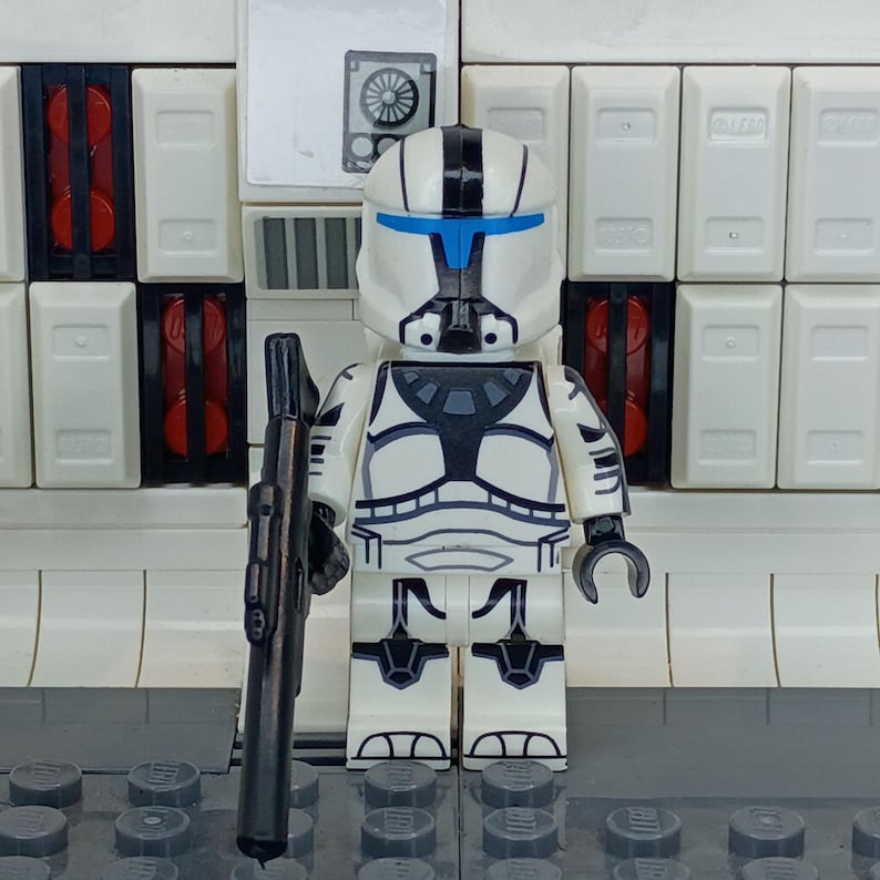 May include: A white and black Lego figure of a clone trooper, holding a black blaster rifle. The figure has blue accents on the helmet and chest. The background features white panels and red accents.