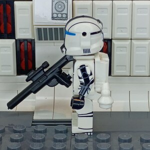 May include: A white and black action figure of a soldier with a black rifle. The figure has a white helmet with blue accents and black markings on the arms and legs. The background features white panels and gray bricks.