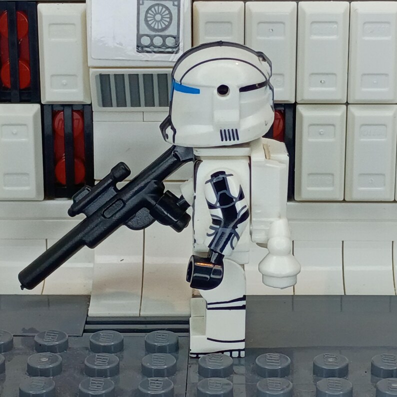 May include: A white and black action figure of a soldier with a black rifle. The figure has a white helmet with blue accents and black markings on the arms and legs. The background features white panels and gray bricks.
