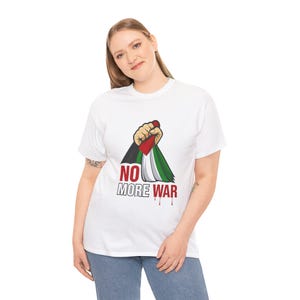 May include: White t-shirt featuring a graphic design with a fist holding a flag with red, white, green, and black colors. The text "NO MORE WAR" is printed below the flag in red, with a dripping effect.