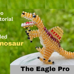 May include: A beaded dinosaur sculpture in shades of gold, orange, and red, with black accents. The dinosaur is posed on a white surface against a blurred green background. Text on the image reads "Video Tutorial Beaded Dinosaur The Eagle Pro."