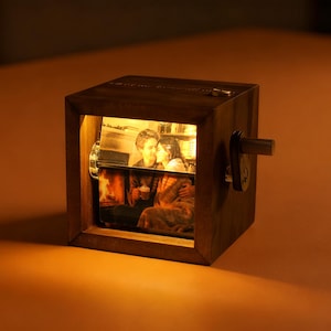 May include: A wooden photo cube with a warm, amber glow illuminating a couple. The cube has a crank and a small door. The image inside shows a couple embracing near a fireplace. The cube is on an orange surface.