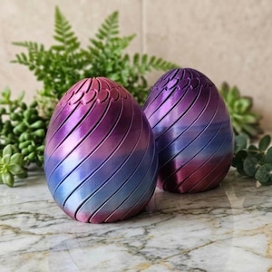 May include: Two decorative, egg-shaped objects with a gradient color scheme of blue, purple, and pink. The eggs have a spiral design, with each layer of color wrapping around the form. The objects are placed on a marble surface.