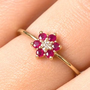 May include: A gold ring featuring a flower-shaped cluster of six round, deep pink rubies surrounding a single, small, clear white diamond at the center. The band is thin, polished gold, and the setting holds the stones in a floral arrangement.