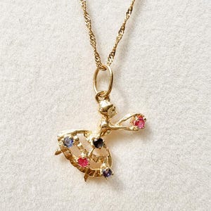 May include: A gold necklace featuring a pendant shaped like a dancing figure. The pendant is adorned with five small, round, multi-coloured gemstones, including pink, blue, and purple stones set into the figure's dress and extended arm.