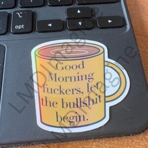 May include: A yellow coffee mug sticker with a gradient orange rim and handle. The text printed on the front of the mug reads, Good Morning fuckers, let the bullshit begin. The sticker is placed on a dark gray computer keyboard with black keys.